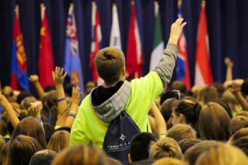 A student stands up raising their hand in a large group of students in front of several flags during Tour Day