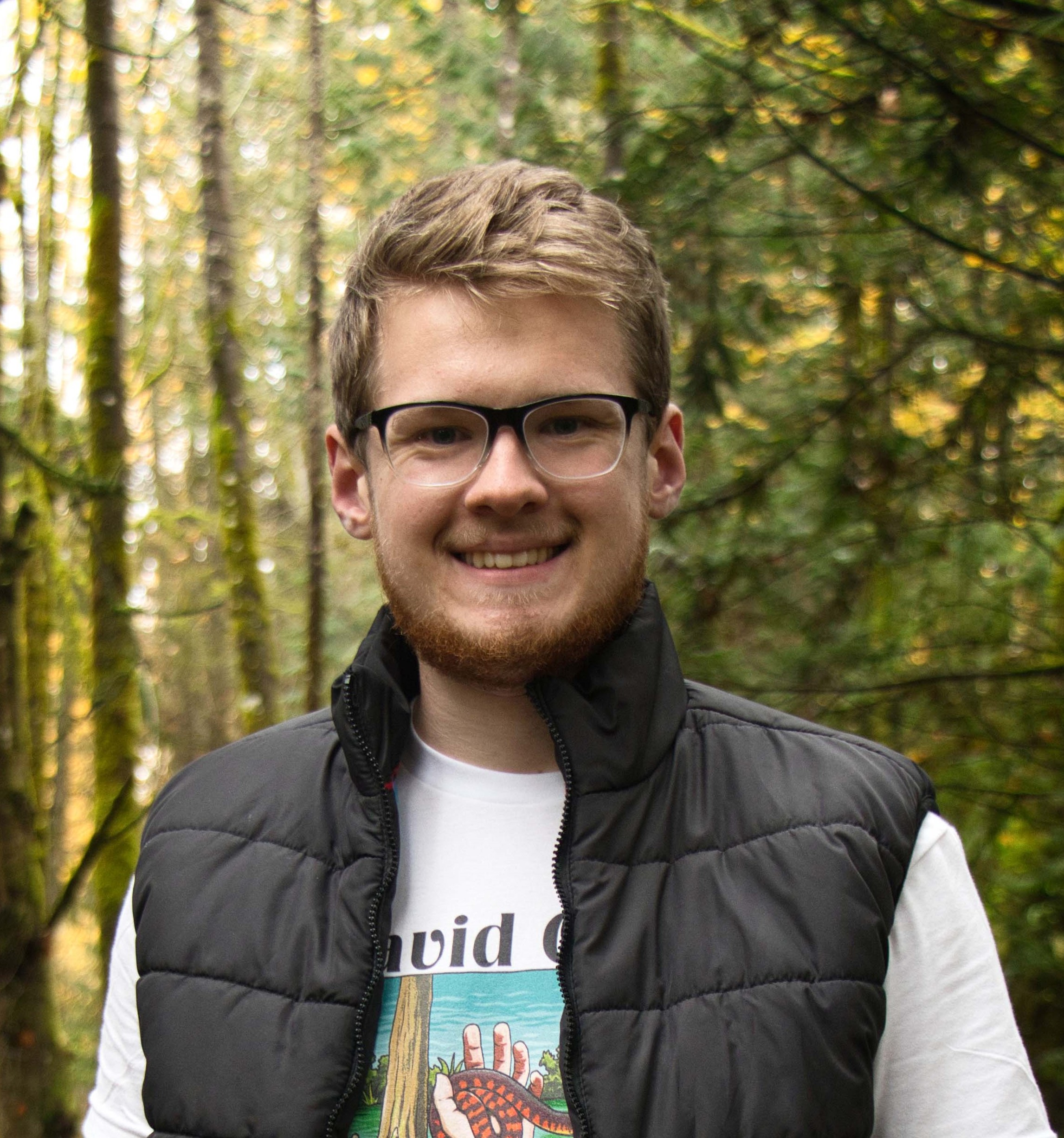 Connor stands in front of trees, with a black vest, white t-shirt and glasses.