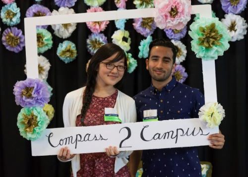 Two staff members smile and hold up a photo booth sign that says Compass 2 Campus with tissue paper flower surrounding them in the background.