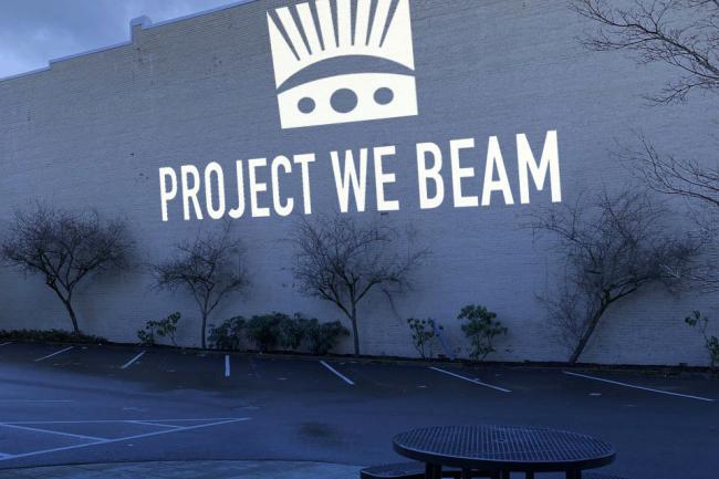 Project We Beam logo and text projected on the side of a building.