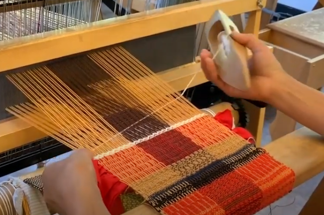 Two hands use a floor loom to weave a piece of fabric that is bright red, navy blue and white