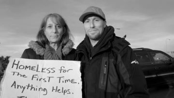 Two people outdoors holding a sign that reads "Homeless for the first time, anything helps"